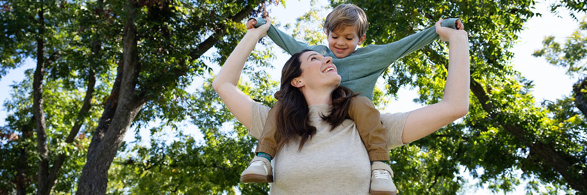 Adult carrying a child on their shoulders while walking outdoors in a park with trees in the background